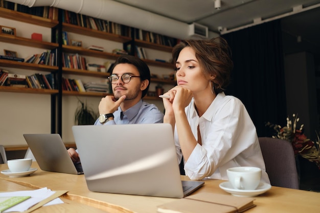 Jovens colegas de negócios sérios trabalhando pensativamente em um novo projeto com laptop e café no escritório moderno