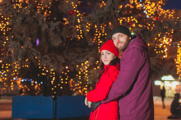 Jovens cônjuges apaixonados posando na rua com decoração de Natal. Jovem de pé atrás de sua esposa, abraçando sua cintura, ambos sorrindo alegremente, olhando para a câmera