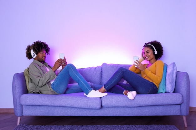 Jovens cônjuges negros usando fones de ouvido ouvindo música juntos em ambientes fechados