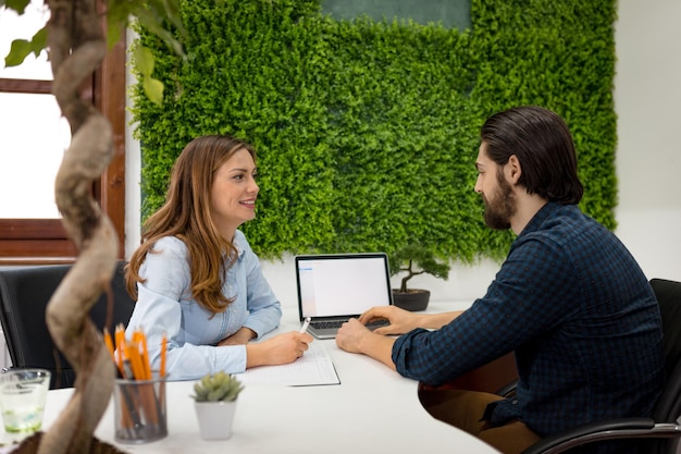 Jovens designers de sucesso falando sobre projeto no escritório. Jovem ouve seu colega e registra os fatos no caderno.