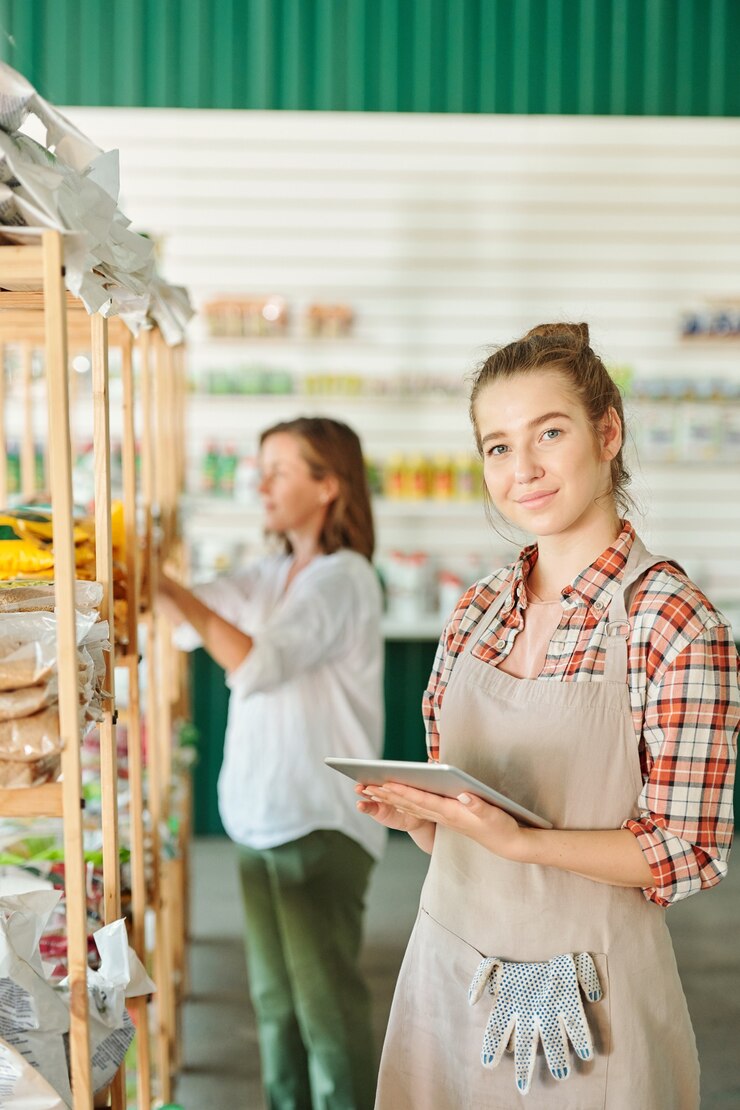 Foto jovens e belas assistente de supermercado de jardinagem em roupas de trabalho usando tablet digital enquanto procuram produtos em sua loja online