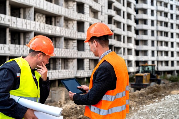 Jovens e fortes trabalhadores da construção civil de macacão estão perto do novo prédio com um plano de trabalho. Conceito de construção