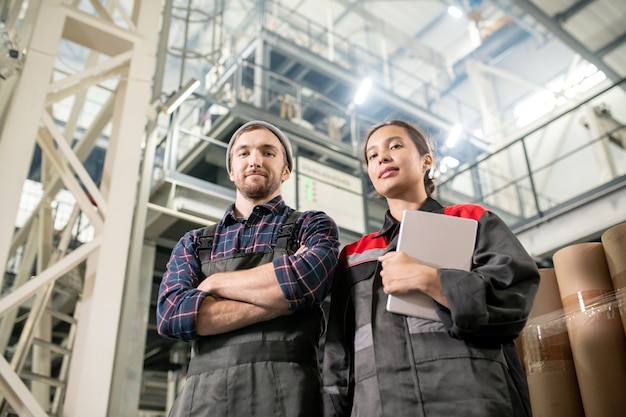 Jovens engenheiros e engenheiros bem-sucedidos de uma grande fábrica em pé na frente da câmera e olhando para você contra uma enorme construção técnica