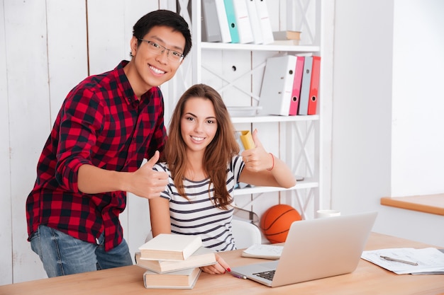 Jovens estudantes felizes olhando para a frente e fazendo gestos de polegar para cima enquanto sorriem