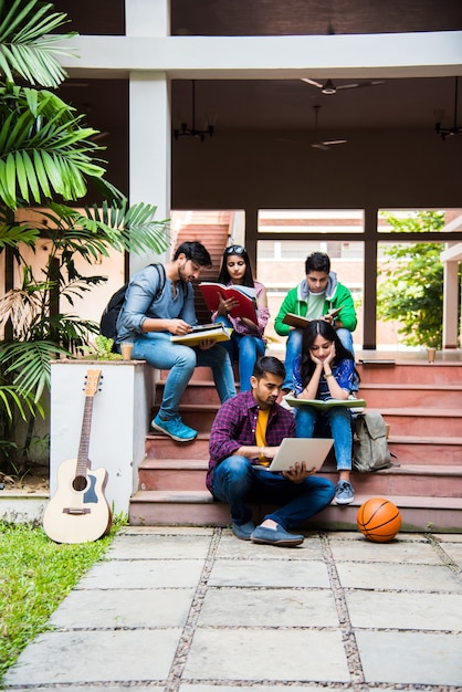 Jovens estudantes universitários indianos asiáticos lendo livros, estudando em um laptop, preparando-se para o exame ou trabalhando em um projeto de grupo enquanto estão sentados na grama, escada ou degraus do campus da faculdade