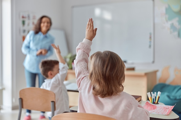 Jovens felizes alunos do ensino fundamental em uma sala de aula moderna estão levantando as mãos para uma lição