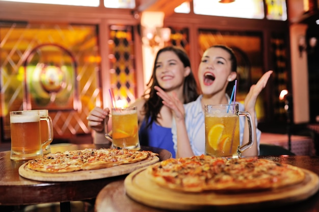 Jovens garotas bonitas comendo pizza, bebendo cerveja ou um coquetel de cerveja e assistindo futebol