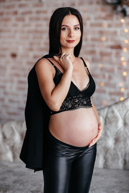 Jovens grávidas de cabelo comprido usando vestido preto e chapéu posando no estúdio Fundo cinza