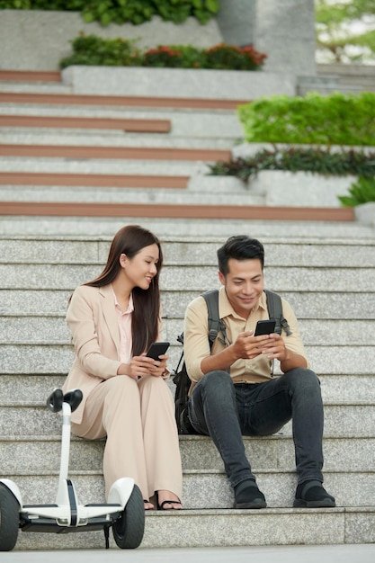 Jovens iniciantes asiáticos sorridentes sentados nos degraus e testando o novo aplicativo móvel que eles criaram