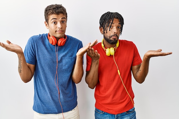 Jovens irmãos hispânicos em pé sobre fundo isolado usando fones de ouvido sem noção e expressão confusa com os braços e as mãos levantadas. conceito de dúvida.