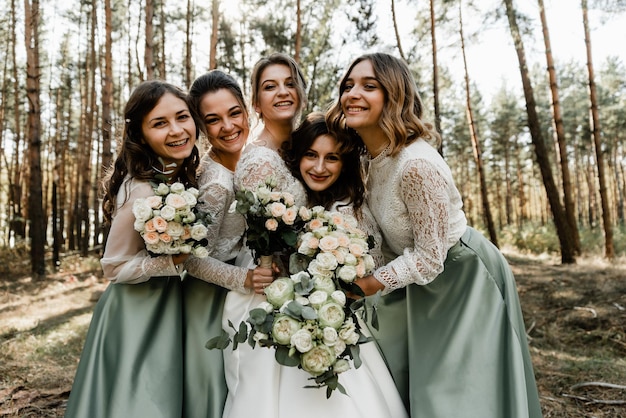 Jovens lindas em vestidos sorrindo e segurando buquês com flores, damas de honra abraçam a noiva com força, companhia alegre, dia do casamento, manhã da noiva