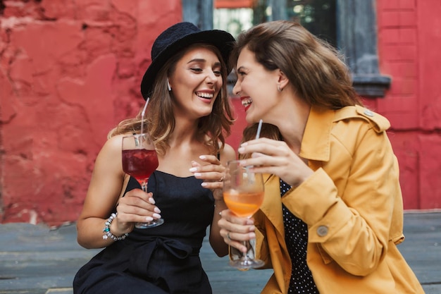 Jovens lindas garotas rindo segurando coquetéis nas mãos alegremente olhando uns para os outros enquanto passam tempo juntos no velho e aconchegante pátio do café