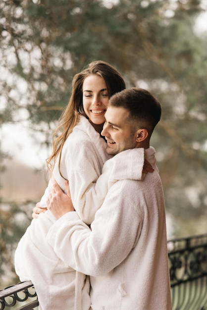 Jovens lindos recém-casados felizes apaixonados por um homem e uma mulher em roupões de banho brancos na varanda da casa sorriem e abraçam