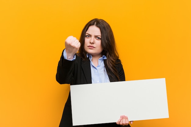 Jovens mais mulher curvilínea de tamanho segurando um cartaz mostrando o punho com expressão facial agressiva.