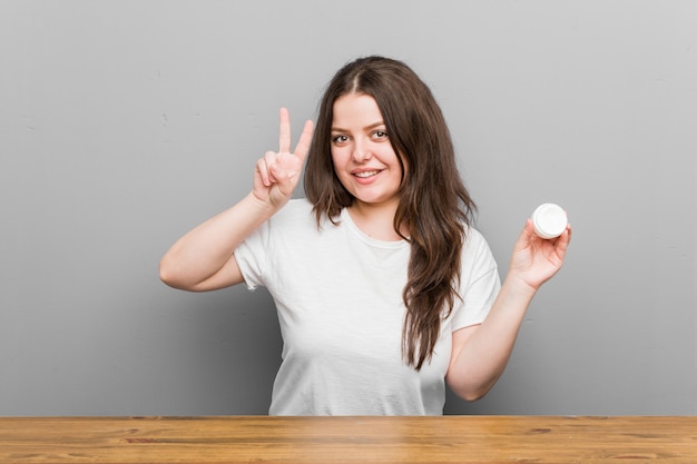 Jovens mais tamanho mulher curvilínea segurando um hidratante mostrando sinal de vitória e sorrindo amplamente.