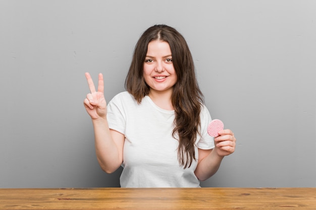 Jovens mais tamanho mulher curvilínea segurando uma esponja facial, mostrando o número dois com os dedos.