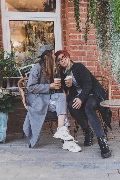 Jovens melhores amigas conversando umas com as outras discutindo temas interessantes durante o tempo livre meninas hipster brincando e se divertindo juntas desfrutando de recreação com café para ir no meio urbano