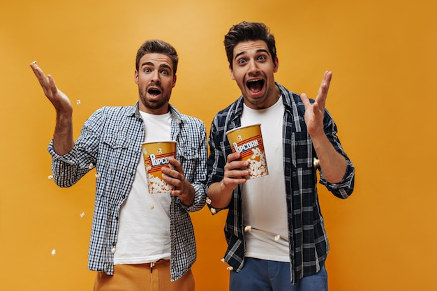 Jovens morenos emocionais loucos em camisetas brancas e camisas xadrez parecem surpresos segurando pipoca e posando em fundo laranja