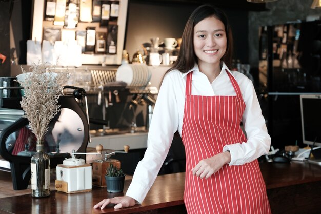 Jovens mulheres asiáticas barista de pé com rosto sorridente na fonte do fundo do contador do café