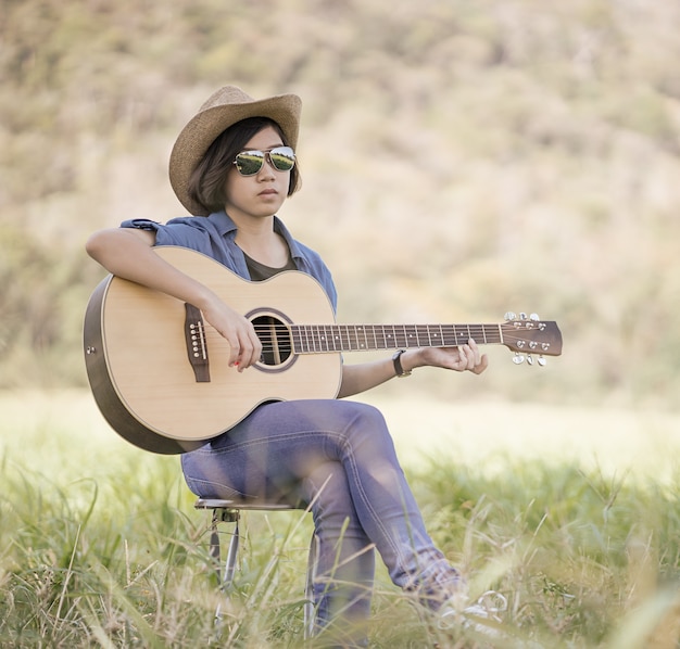 Jovens mulheres asiáticas, cabelos curtos, usam chapéu e óculos de sol, sentam-se tocando guitarra em um campo de grama na Tailândia