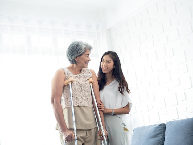 Jovens mulheres asiáticas sorridentes seguram e apoiam com cuidado os braços de mulheres idosas tentando andar de muletas na sala de recuperação, ajudando mulheres idosas a terapia de cuidados de saúde paciente sênior em casa conceito