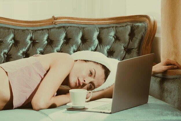 Jovens mulheres bonitas com café e laptop em roupões de banho brancos e toalhas no quarto depois de acordar