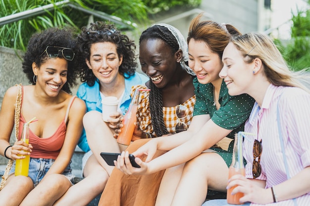 Jovens mulheres diversas se divertindo ao ar livre rindo juntas e usando o aplicativo de smartphone para o conceito de diversidade de mídia social foco principal no rosto da garota africana