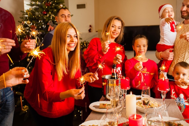 Jovens mulheres sorridentes felizes, homens e crianças, todos em roupas vermelhas, reunidos ao redor da mesa de férias, segurando brilhos iluminados, desejando feliz Ano Novo um para o outro