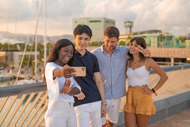 Jovens multirraciais positivos com smartphone tirando selfie juntos durante o encontro em um dique urbano na noite de verão