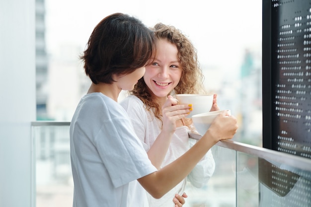Jovens namoradas alegres e apaixonadas em pé na varanda, tomando café da manhã e olhando uma para a outra