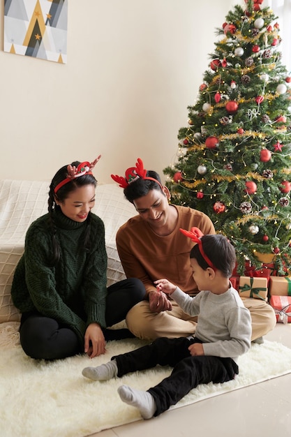 Jovens pais brincando com o filho quando estão sentados no chão em casa ao lado da árvore de Natal