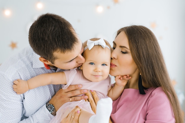 Foto jovens pais felizes seguram sua filha de um ano nos braços e a beijam na bochecha