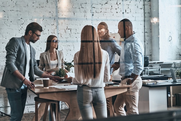 Jovens profissionais de negócios. Grupo de jovens modernos em roupas casuais inteligentes discutindo negócios em pé atrás da parede de vidro no escritório criativo