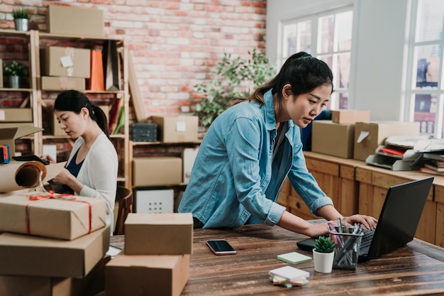 Jovens proprietários do sexo feminino iniciam para o projeto de entrega de PME on-line de negócios. empregado de duas meninas asiáticas concentrado trabalhando em laptop e tablet pronto para enviar produtos de caixas no escritório