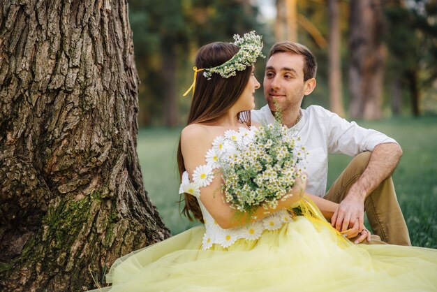 Jovens recém-casados felizes sentados na grama perto de uma árvore no parque