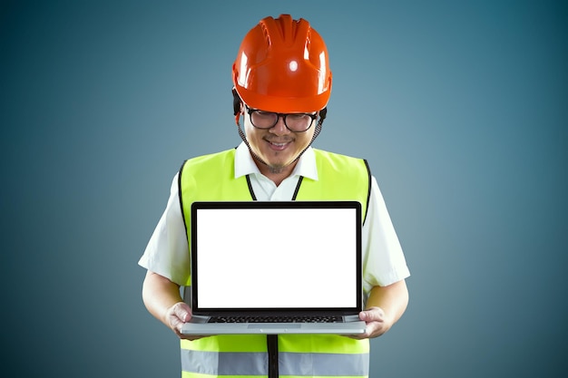 Jovens trabalhadores da construção civil asiáticos em capacete de segurança e usando laptop com tela vazia para mostrar ao cliente algo isolado em fundo azul