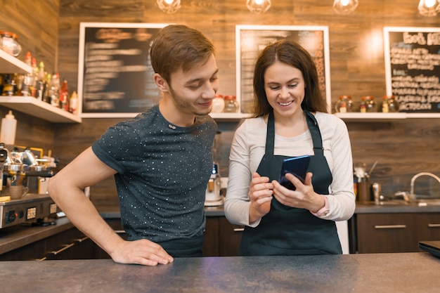 Jovens trabalhadores de café homem e mulher atrás do balcão de bar, falando olhando para smartphone