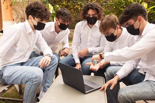 Foto jovens usando máscara usando desinfetante de mãos enquanto estão sentados ao lado do laptop ao ar livre