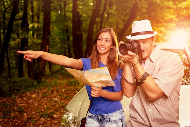 Foto jovens viajantes em pé diante de um carro na floresta. menina segurando o mapa e apontando o destino. cara tirando uma foto com câmera digital.