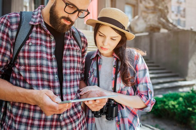 Jovens viajantes ficam lado a lado e olham para o tablet. O homem mantém isso no hads. Eles são sérios e concentrados. Os turistas usam mochilas nas costas.