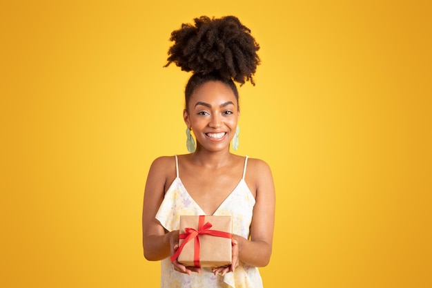 Jovial joven negra sosteniendo una caja con un regalo aislado sobre un fondo amarillo