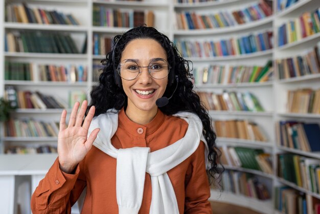 Foto joyful curly garota hispânica sentada em uma mesa na frente de um laptop em um fone de ouvido olhando para o