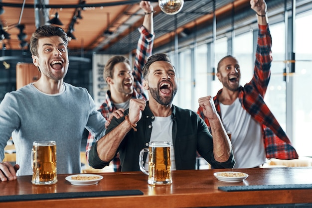 Jubelnde junge Männer in Freizeitkleidung trinken Bier und beobachten Sportspiele, während sie in der Kneipe sitzen