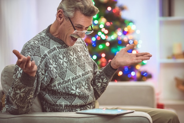 El jubilado feliz en anteojos con una tableta se sienta cerca del árbol de Navidad