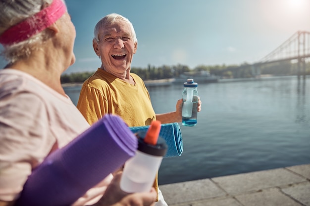 Jubilado masculino animado con una estera de yoga y una botella de deportes mirando a una mujer