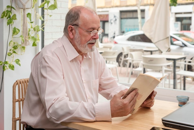 Un jubilado mayor feliz disfrutando de leer un libro sentado en una mesa de madera en una cafetería