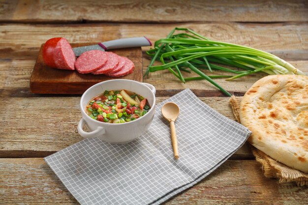 Jüdische Bohnensuppe mit geräuchertem Fleisch in einer Terrine auf einer grauen Serviette neben einer Wurst und einem Fladenbrot