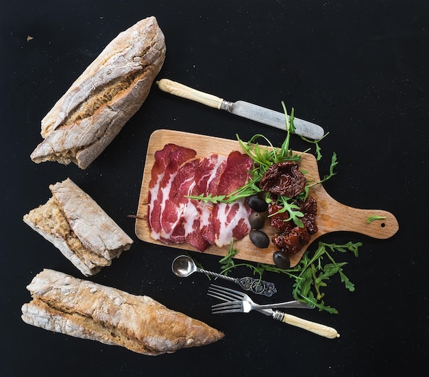 Juego de aperitivos de vino vajilla vintage baguette francesa rota en pedazos tomates secos aceitunas carne ahumada y rúcula en una tabla de madera rústica sobre fondo oscuro