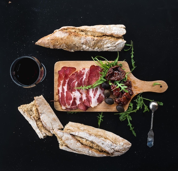 Juego de aperitivos de vino vajilla vintage baguette francesa rota en pedazos tomates secos aceitunas carne ahumada y rúcula en una tabla de madera rústica sobre fondo oscuro