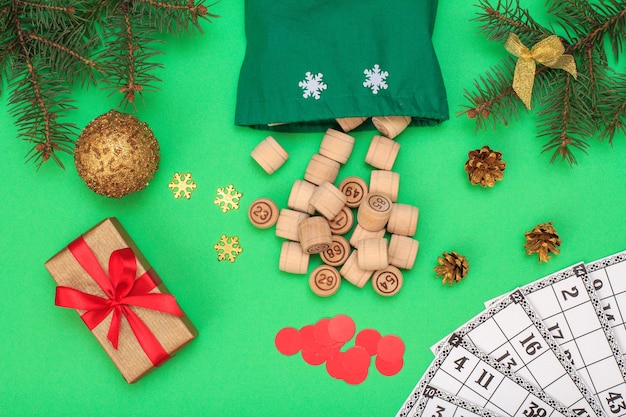 Foto juego de mesa de lotería. barriles de madera con bolsa, tarjetas de juego y fichas rojas para un juego de lotería, rama de abeto de navidad, cono, pelota de juguete y cajas de regalo sobre fondo verde. vista superior